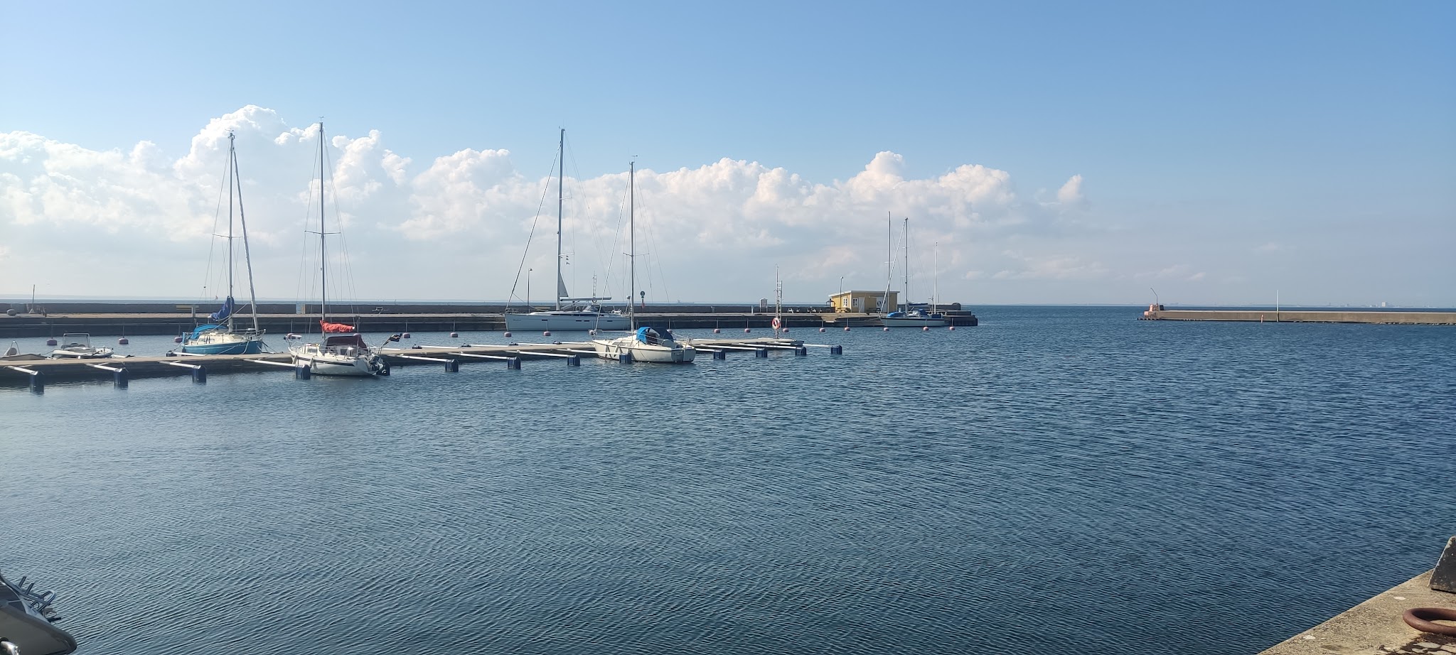 Ruhiger Yachthafen; mehrere Segelboote liegen an Stegen, dahinter Wellenbrecher, blauer Himmel mit leichten Wolken.