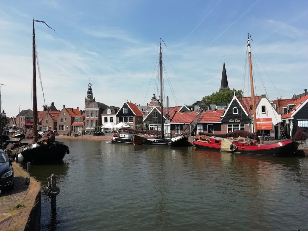 Historischer niederländischer Hafen mit roten Giebelhäusern, Kirchturm und mehreren Traditionsseglern im Wasser.