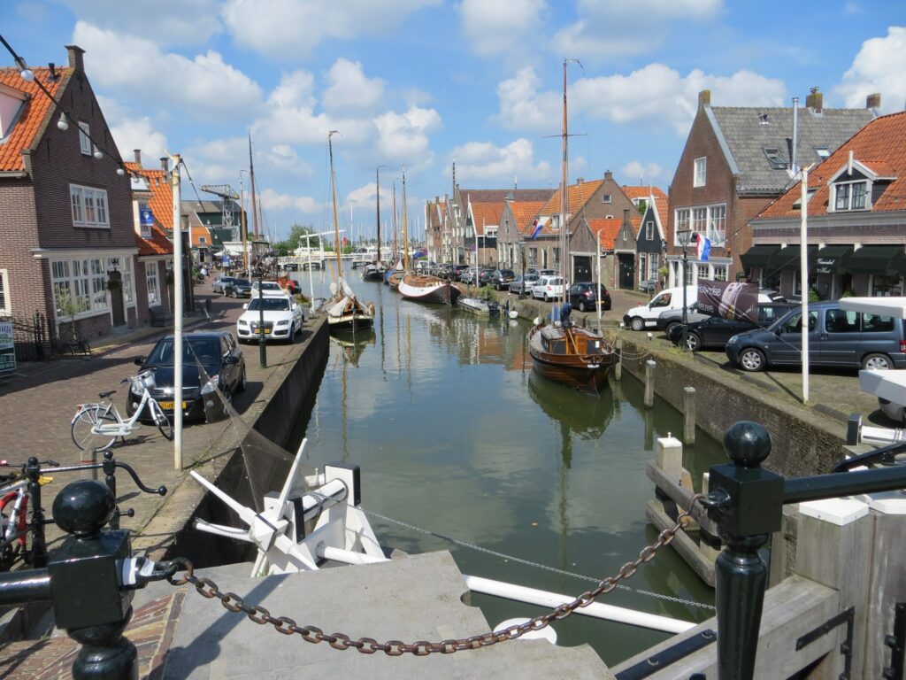 Schmaler niederländischer Kanal mit Segelbooten, Backsteinhäusern und parkenden Autos unter blauem Himmel.
