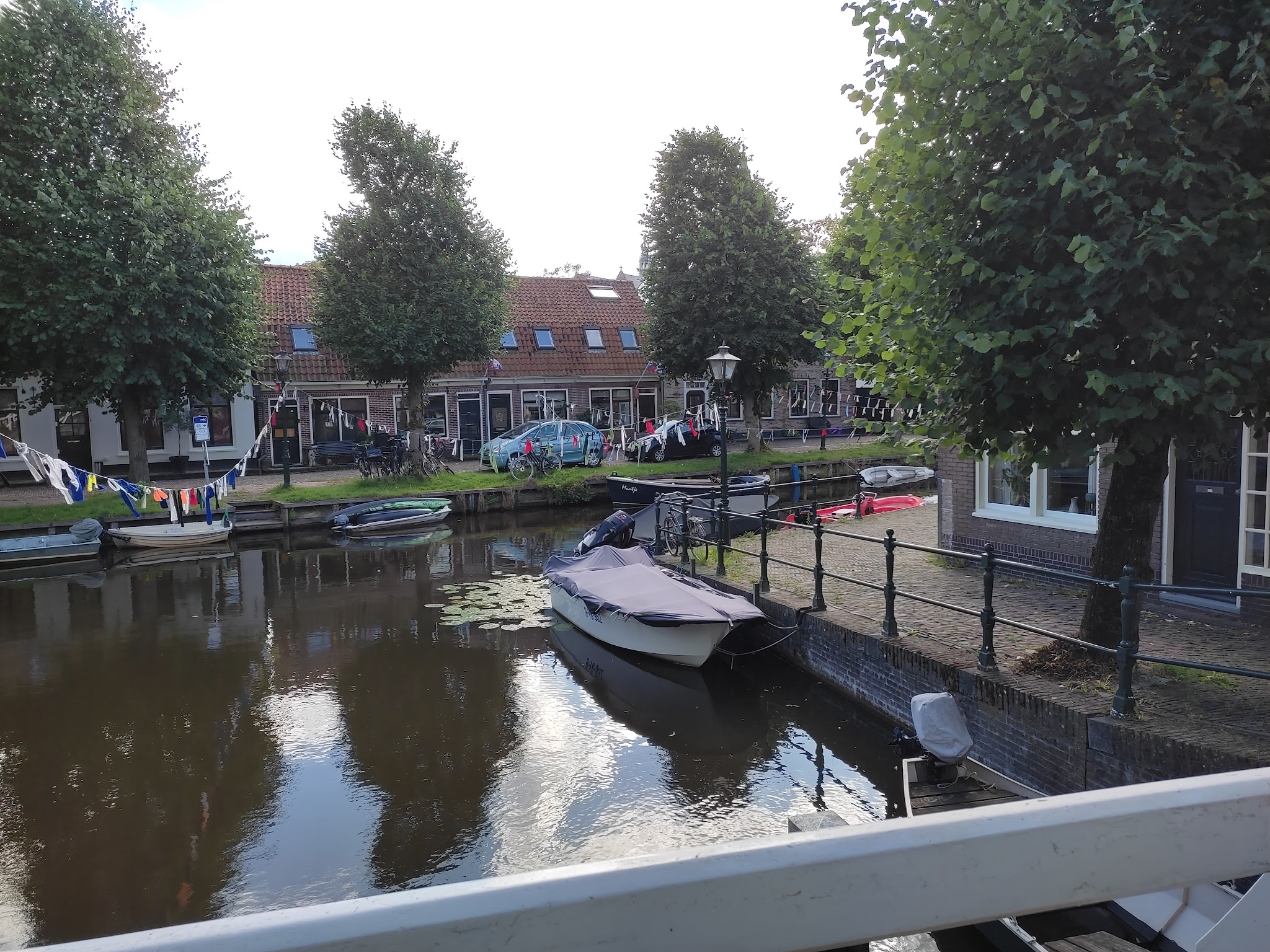 Kleiner Kanal mit vertäuten Booten, bunte Wimpelketten am Ufer vor Backsteinhäusern und schattigen Bäumen.