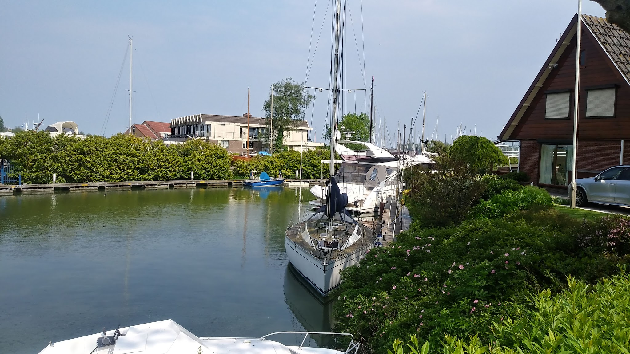 Segel- und Motorboote liegen in schmalem, grünen Hafenkanal neben Haus und Hecken unter blauem Himmel.