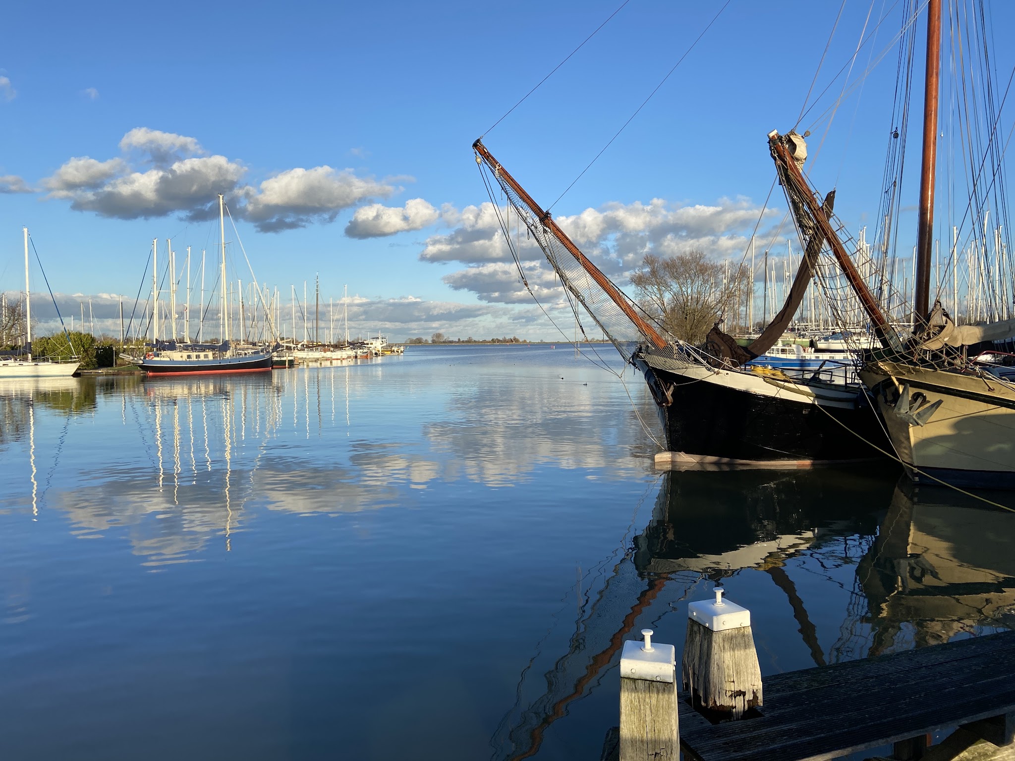 Segelboote liegen in ruhigem Hafen; blauer Himmel und Wolken spiegeln sich im stillen Wasser.