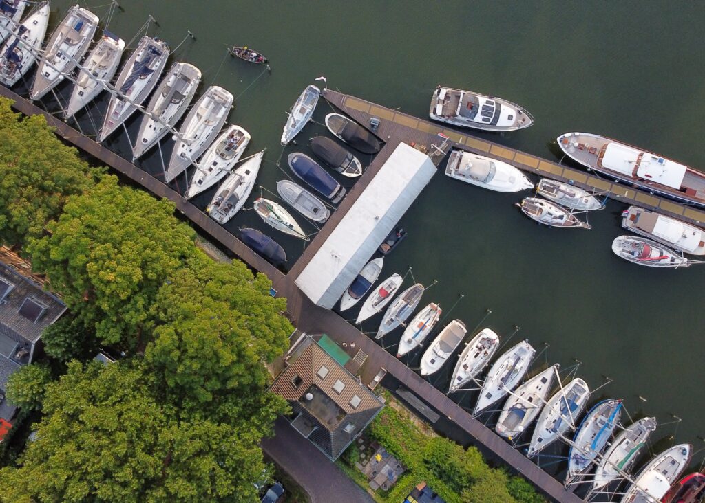 Drohnenansicht eines kleinen Yachthafens: Reihen von Segel- und Motorbooten an Stegen; links dichter Baumbestand