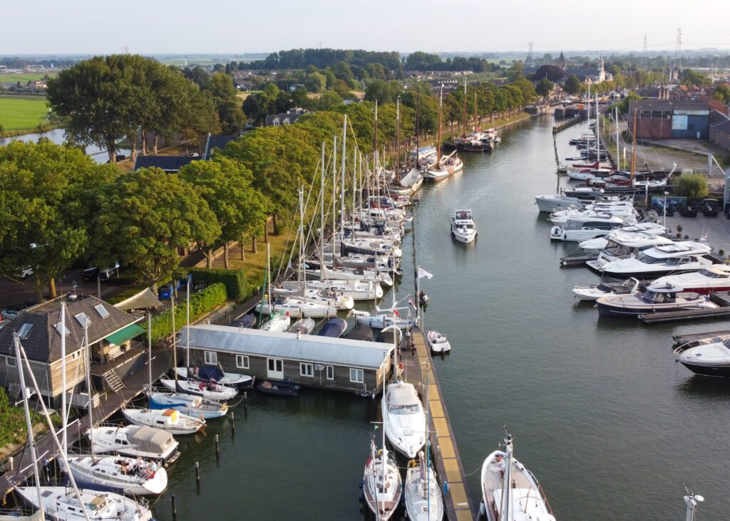 Yachthafen an einem Kanal; Reihen von Segel- und Motorbooten, baumbestandene Promenade, Dorf im Hintergrund