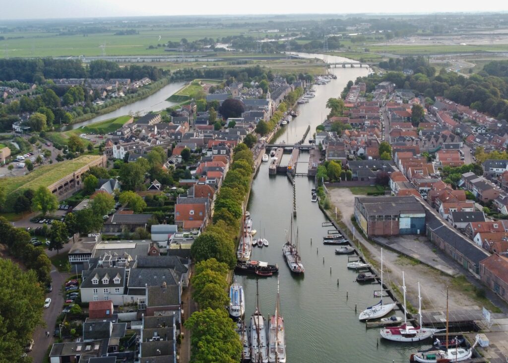Luftaufnahme: schmaler Kanal mit Segelbooten durch kleinstädtische Backsteinhäuser, umgeben von Wiesen und Bäumen.