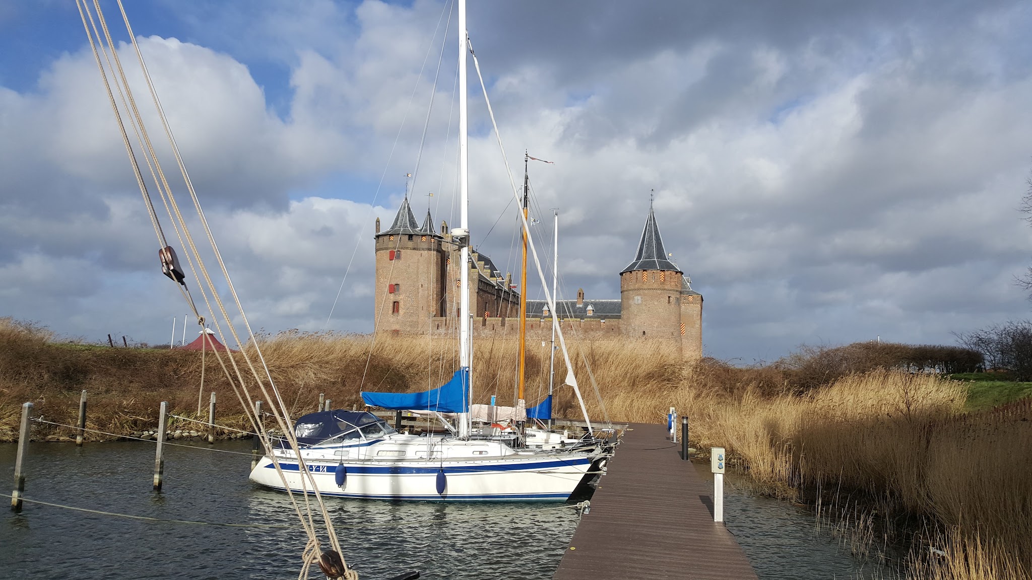 Segelboot am Holzsteg vor mittelalterlicher Burg, umgeben von Schilf, unter teils bewölktem Himmel