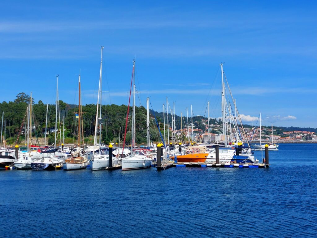 Segelboote im Yachthafen vor blauem Meer; grüner Hügel und Küstenstadt unter wolkenlosem Himmel im Hintergrund.