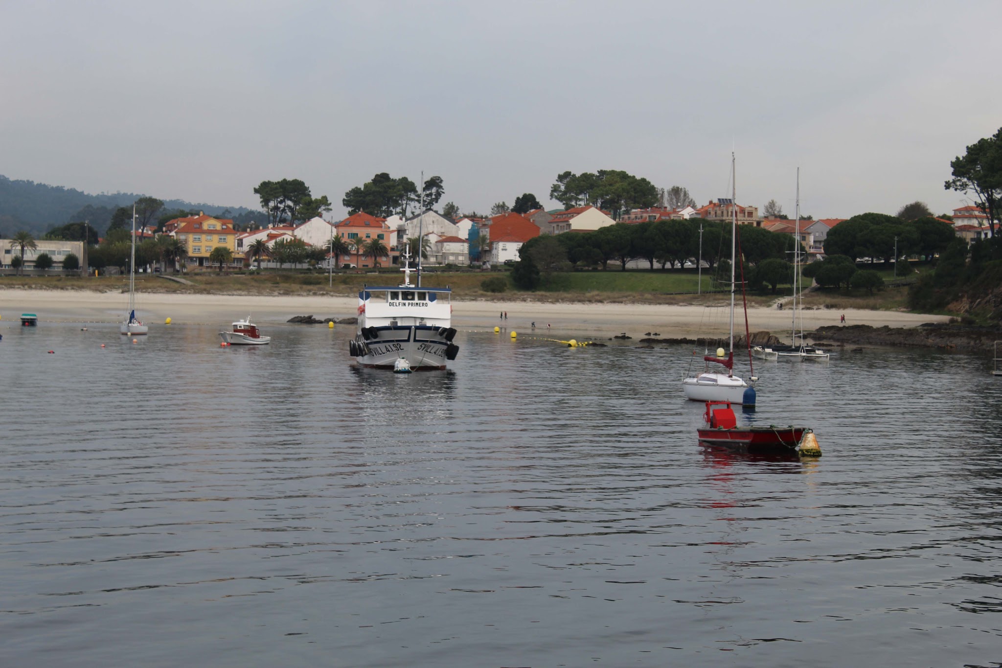 Mehrere Boote, darunter eine kleine Fähre, liegen vor einem ruhigen Strand mit bunten Häusern und Pinien.