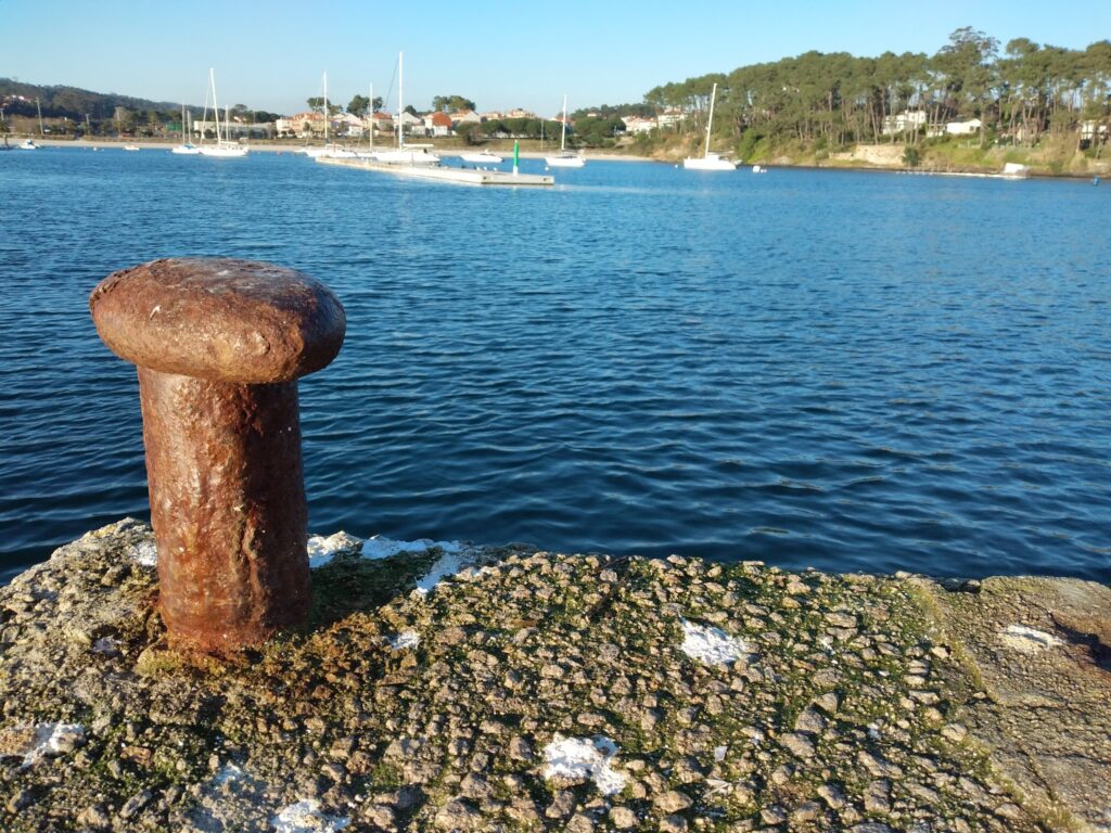 Verrosteter Poller auf Kai, dahinter ruhiges Hafenwasser mit mehreren Segelbooten und bewaldetem Ufer.