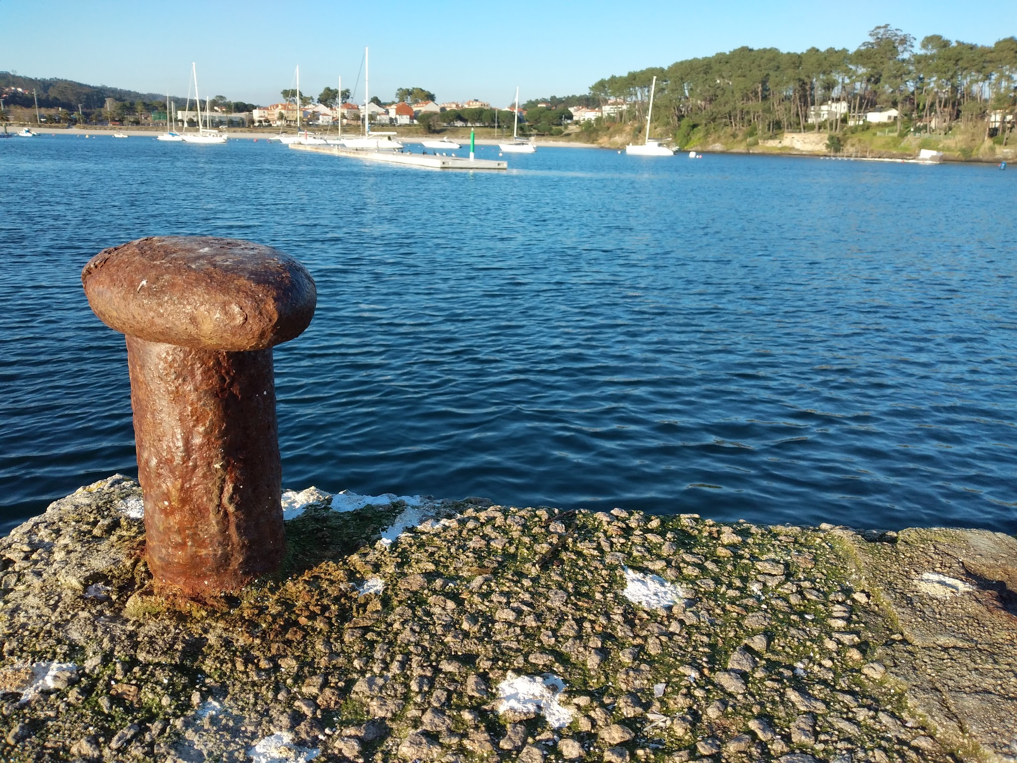 Verrosteter Poller auf Kai, dahinter ruhiges Hafenwasser mit mehreren Segelbooten und bewaldetem Ufer.