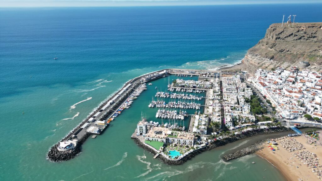 Luftaufnahme eines Yachthafens voller Segelboote, flankiert von weißem Küstenort, Strand und türkisblauem Meer.