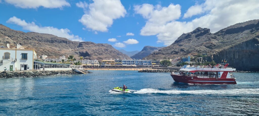 Blaues Meer; Jet-Ski und rotes Ausflugsboot vor Sandstrand, weißer Küstenstadt und kahlen Bergen unter blauem Himmel.