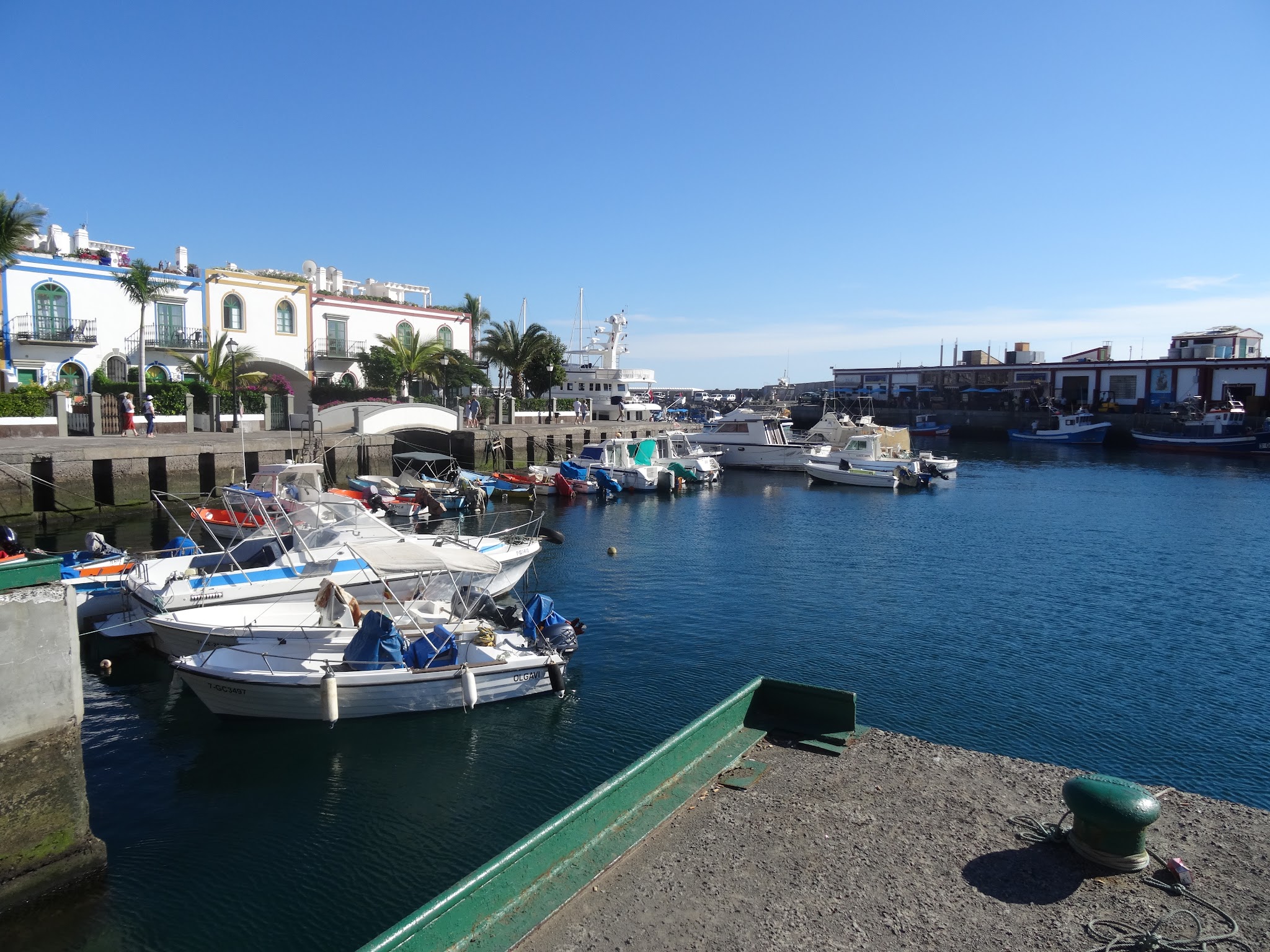 Kleiner Hafen mit weißen Häusern, Palmen und mehreren vertäuten Booten im klaren Wasser unter blauem Himmel.