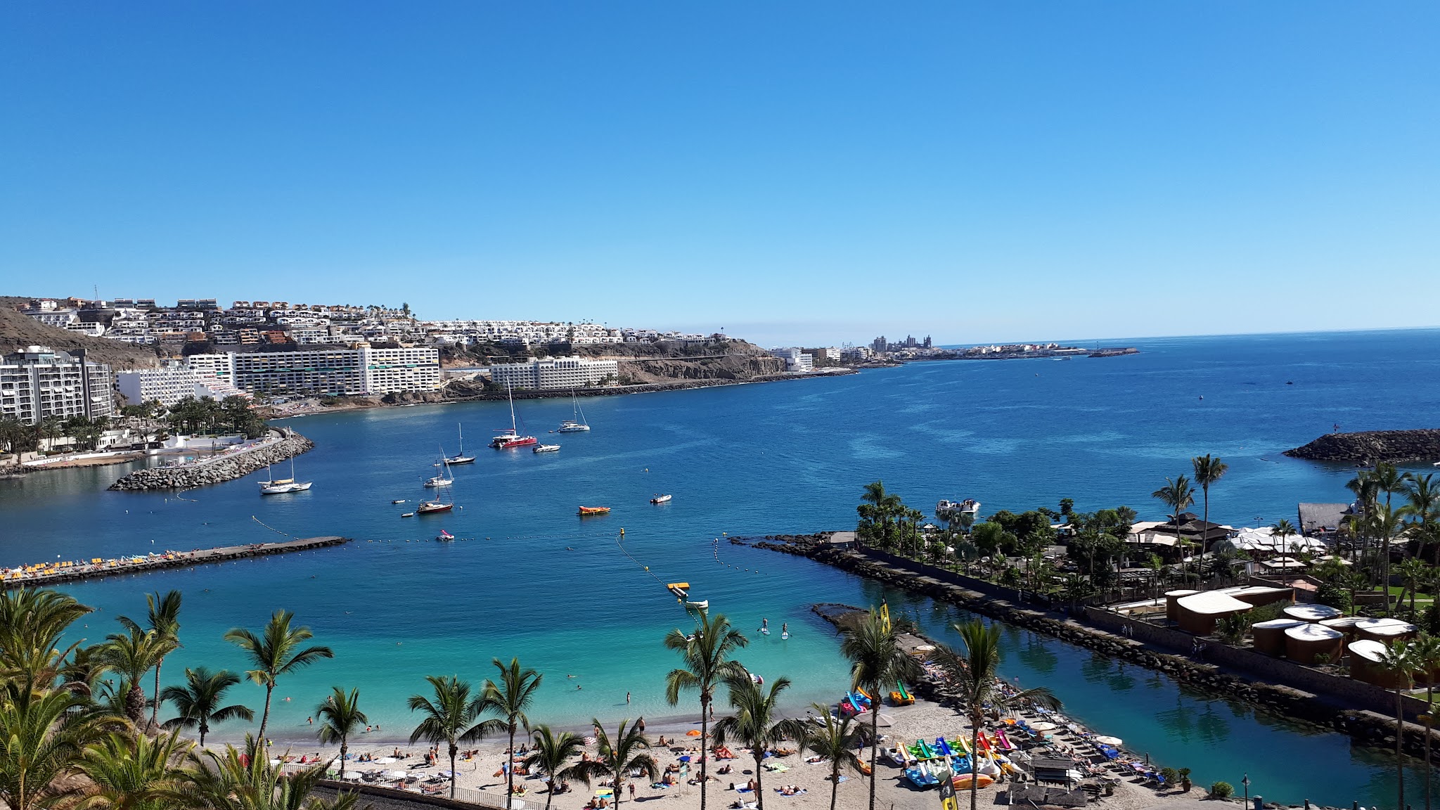 Palmenstrand an türkisblauer Bucht, Badende im Wasser, bunte Liegen, Segelboote vor terrassenförmigen Hotels