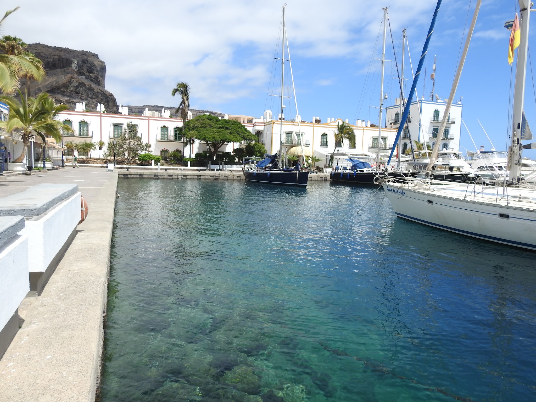 Türkises Hafenbecken mit Segelbooten; weiße Häuser, Palmen und Felswand im Hintergrund.