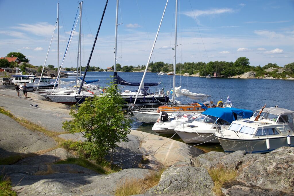 Segel- und Motorboote liegen an felsiger Schärenküste im Sonnenschein; blaue See und vereinzelte Spaziergänger