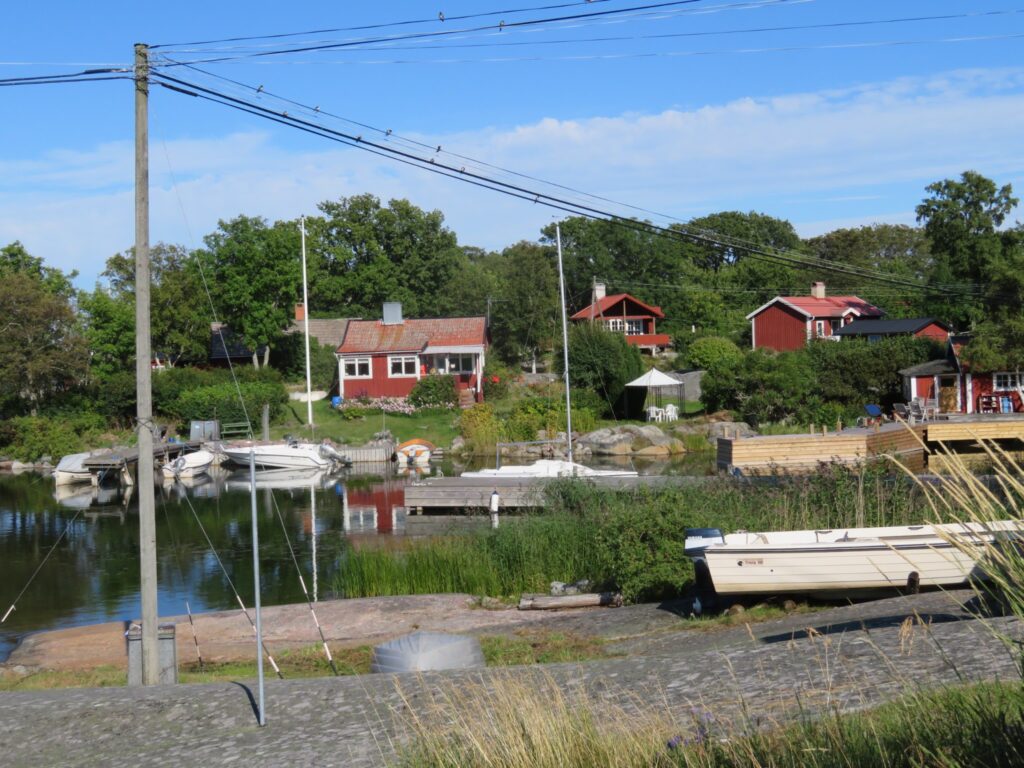 Kleine Motorboote liegen an ruhiger Bucht vor roten Holzhäusern und grüner Landschaft im Sommer
