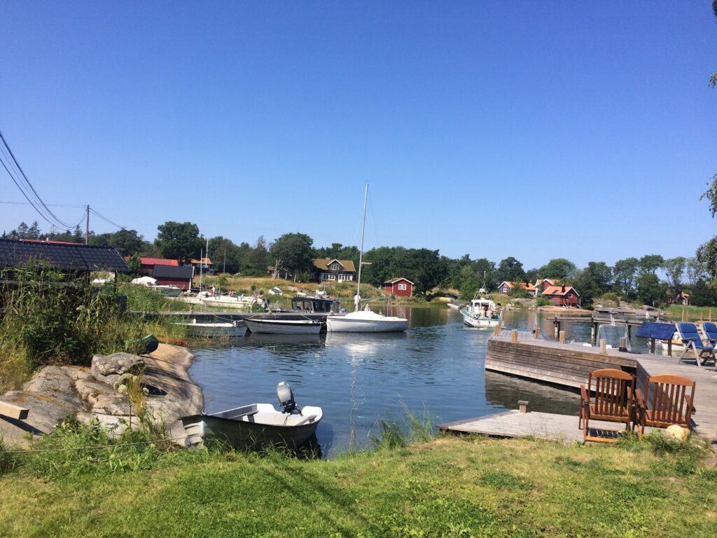 Sonniger kleiner Hafen mit Segel- und Motorbooten, roten Holzhäusern am Ufer, Steg mit zwei Holzstühlen im Vordergrund