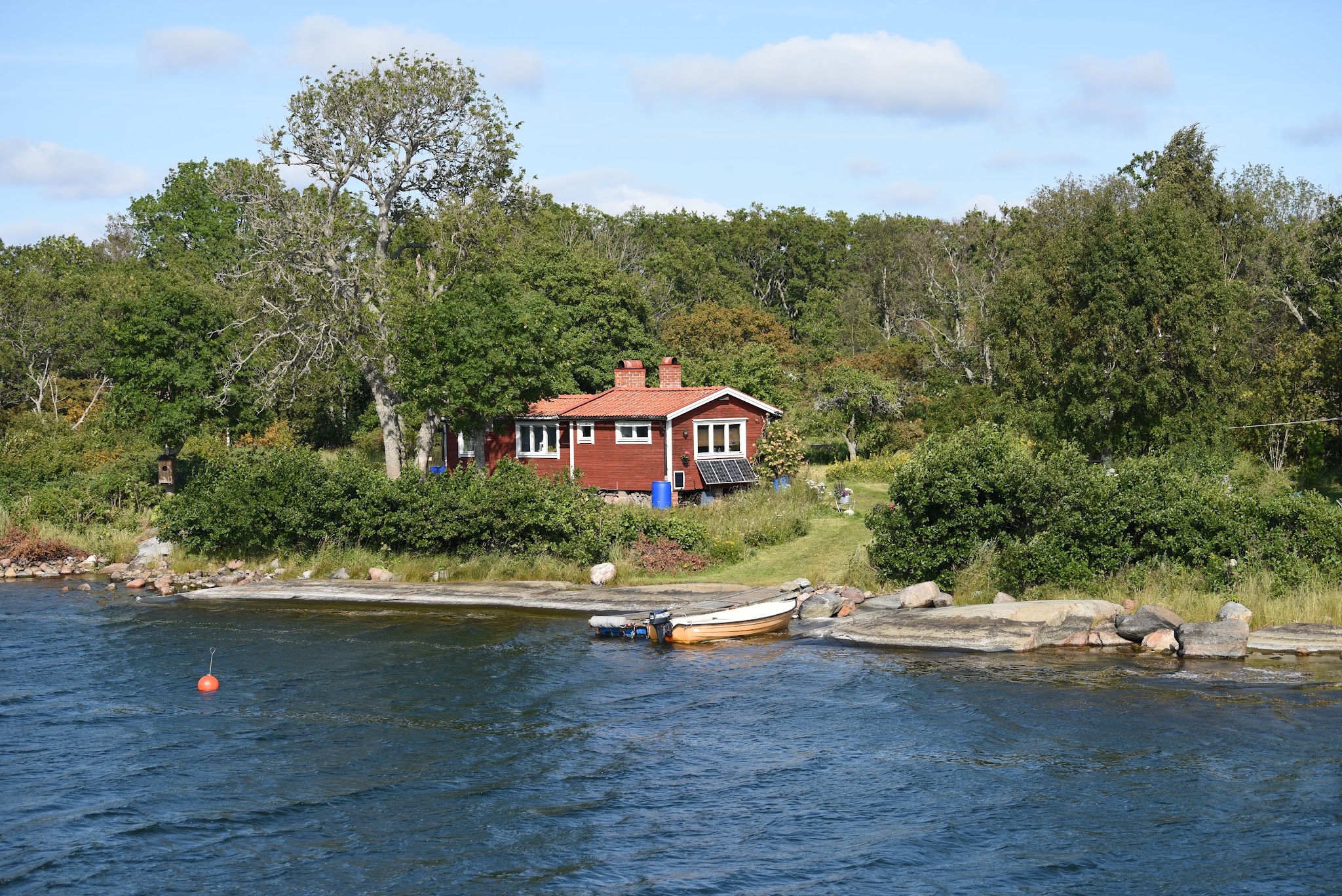 Rotes Holzhaus am bewaldeten Ufer, kleines Boot an Felsen im blauen See unter leicht bewölktem Himmel.