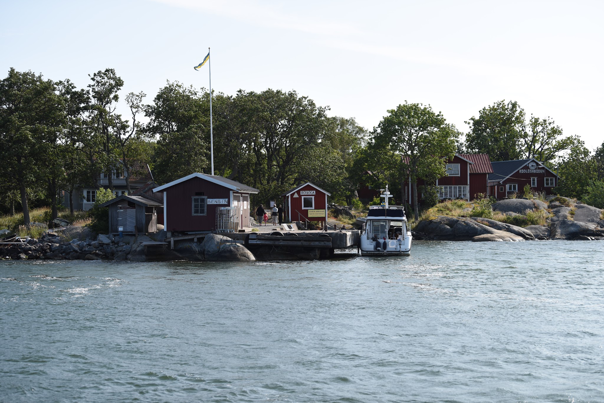 Kleiner Anleger mit roten Holzhäusern, schwedischer Flagge und anlegendem Motorboot, umgeben von Wasser und Bäumen.
