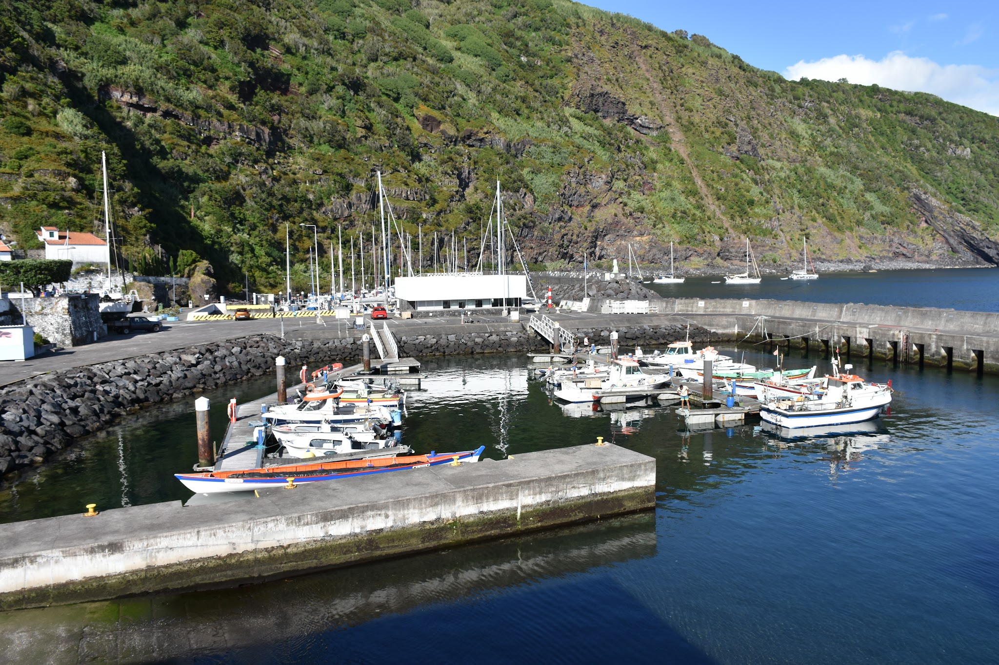 Kleine Marina mit Fischerbooten vor steil bewaldetem Vulkanhang, ruhiges blaues Wasser bei Sonnenschein.