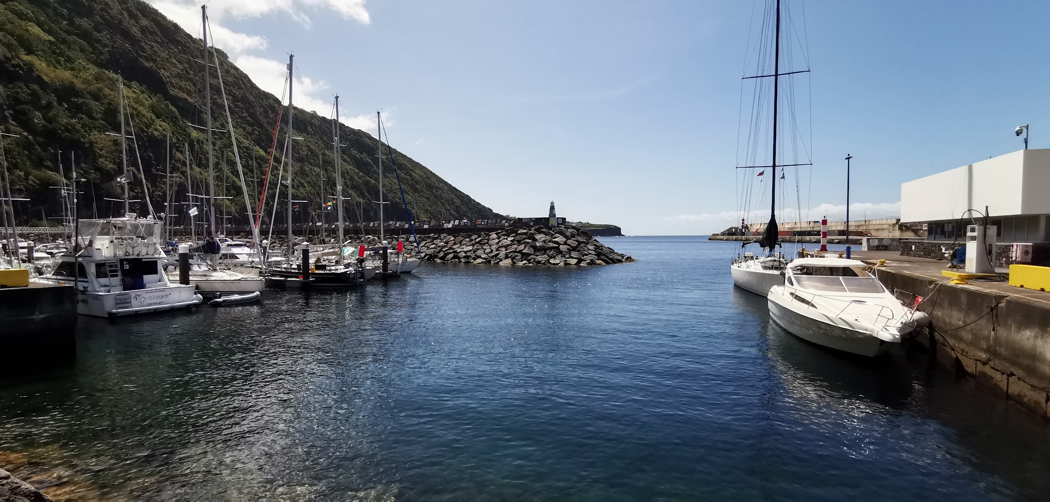 Sonnige Marina: Segelboote vor grünem Steilhang; Felsmole mit kleinem Leuchtturm öffnet sich zum Meer.