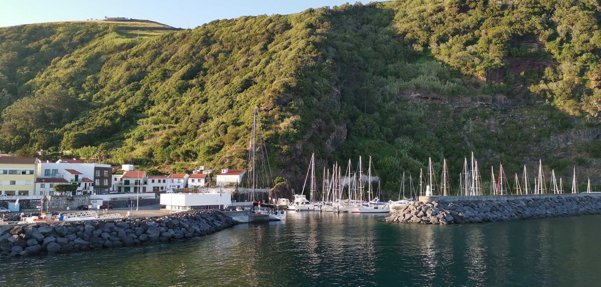 Kleiner Hafen mit weißen Häusern und roten Dächern am grünen Steilhang; Segelboote spiegeln sich im ruhigen Wasser.