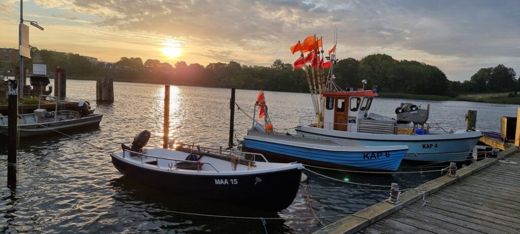 Zwei Fischerboote mit roten Fähnchen vertäut am Steg, Sonnenuntergang spiegelt sich im ruhigen Flusswasser.