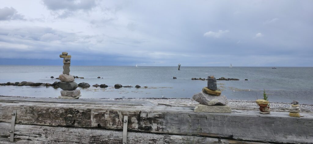 Mehrere Steinmännchen auf Holzplanke vor ruhiger Küste; dahinter Felsen, graublaues Meer und Segelboote unterm Wolkenhimmel