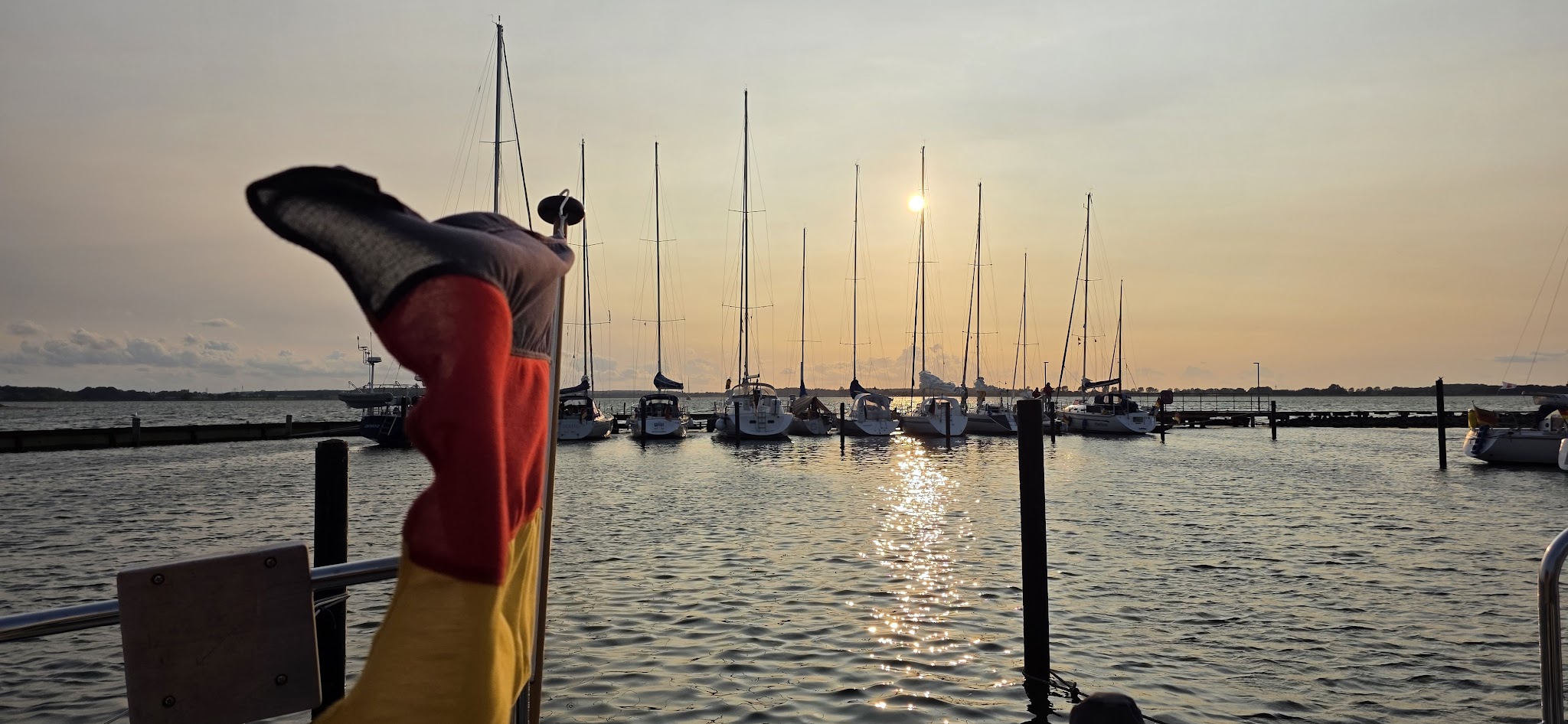 Deutsche Flagge weht im Vordergrund, dahinter liegen Segelboote im Hafen, Sonne spiegelt sich im ruhigen Abendwasser.