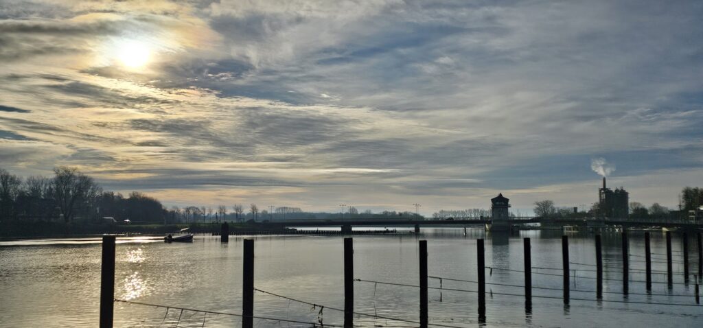 Grelles Morgenlicht über ruhigem Fluss; Pfähle vorn, Brücke, Wachhäuschen und rauchender Schornstein am Horizont
