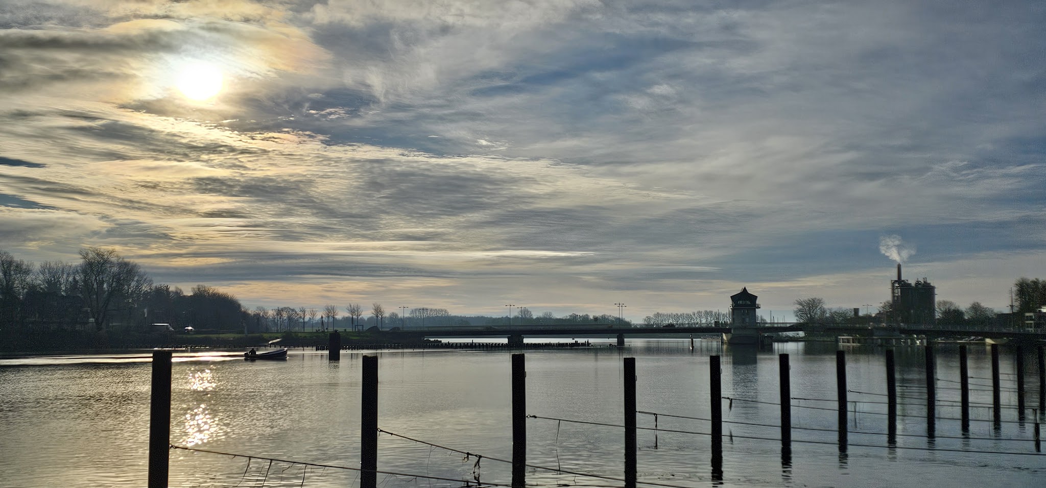 Grelles Morgenlicht über ruhigem Fluss; Pfähle vorn, Brücke, Wachhäuschen und rauchender Schornstein am Horizont
