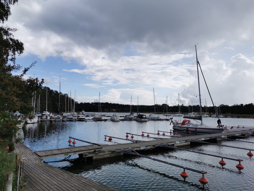 Marina mit Holzstegen, vertäuten Segelbooten, einem Mann am Motorboot und Wolken über ruhigem Wasser.