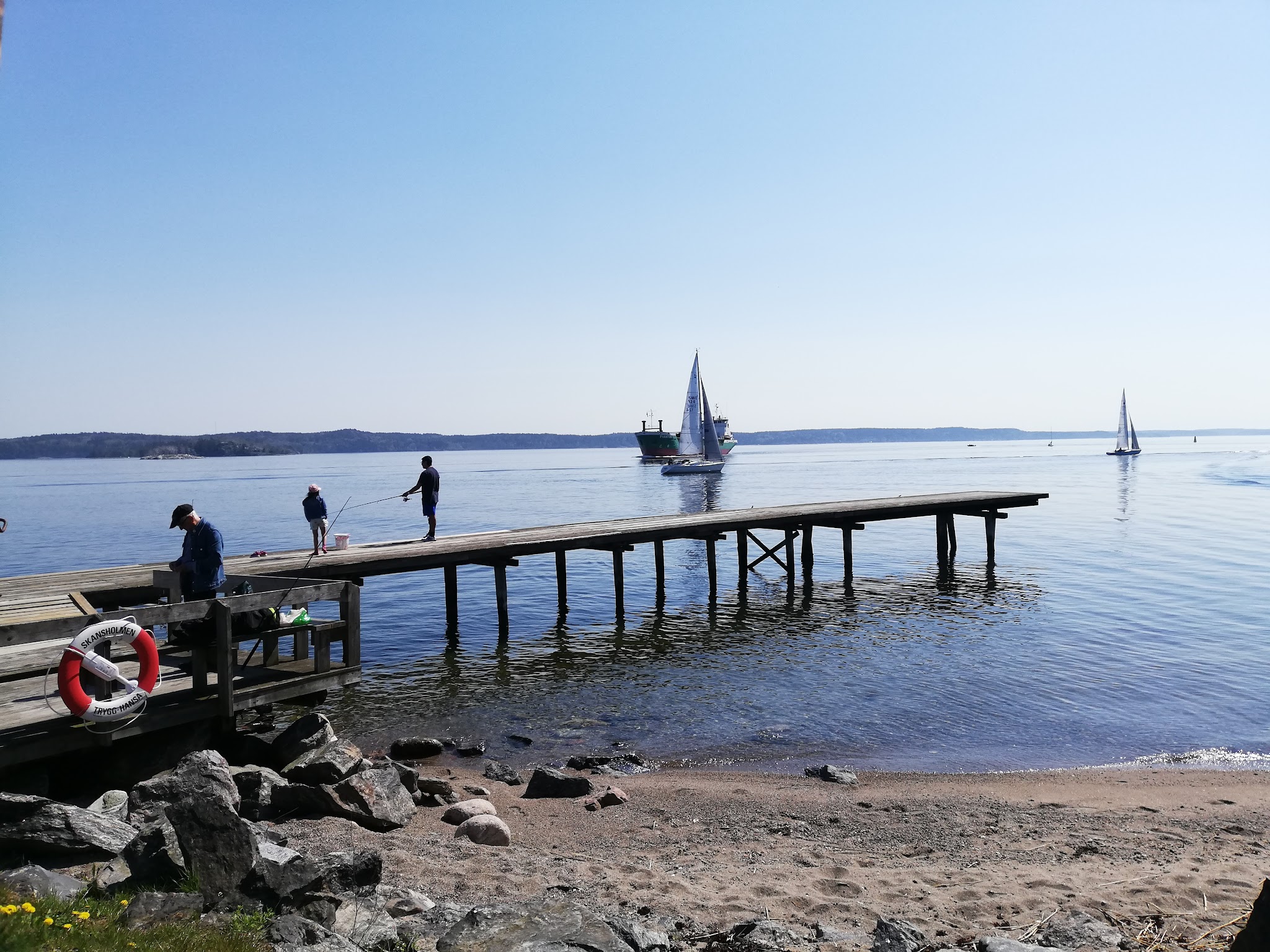 Holzsteg an ruhiger Bucht, drei Personen angeln, dahinter zwei Segelboote auf spiegelglattem Wasser unter blauem Himmel