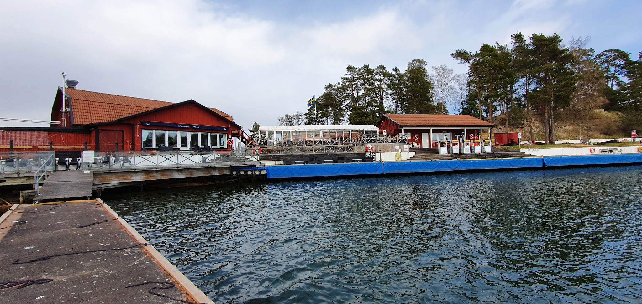 Holzrestaurant in roten Häusern am Steg, blau verkleideter Schwimmsteg auf ruhigem Wasser, schwedische Flagge weht.