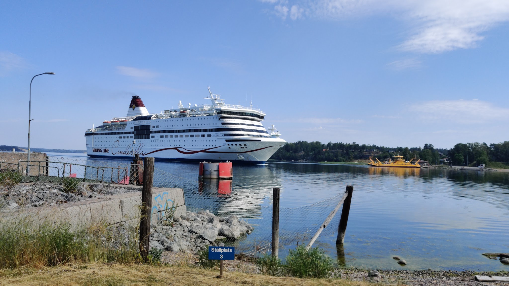 Große weiße Fähre Viking Line Cinderella fährt ruhig übers Wasser; hinten kleinere gelbe Fähre und Waldküste.