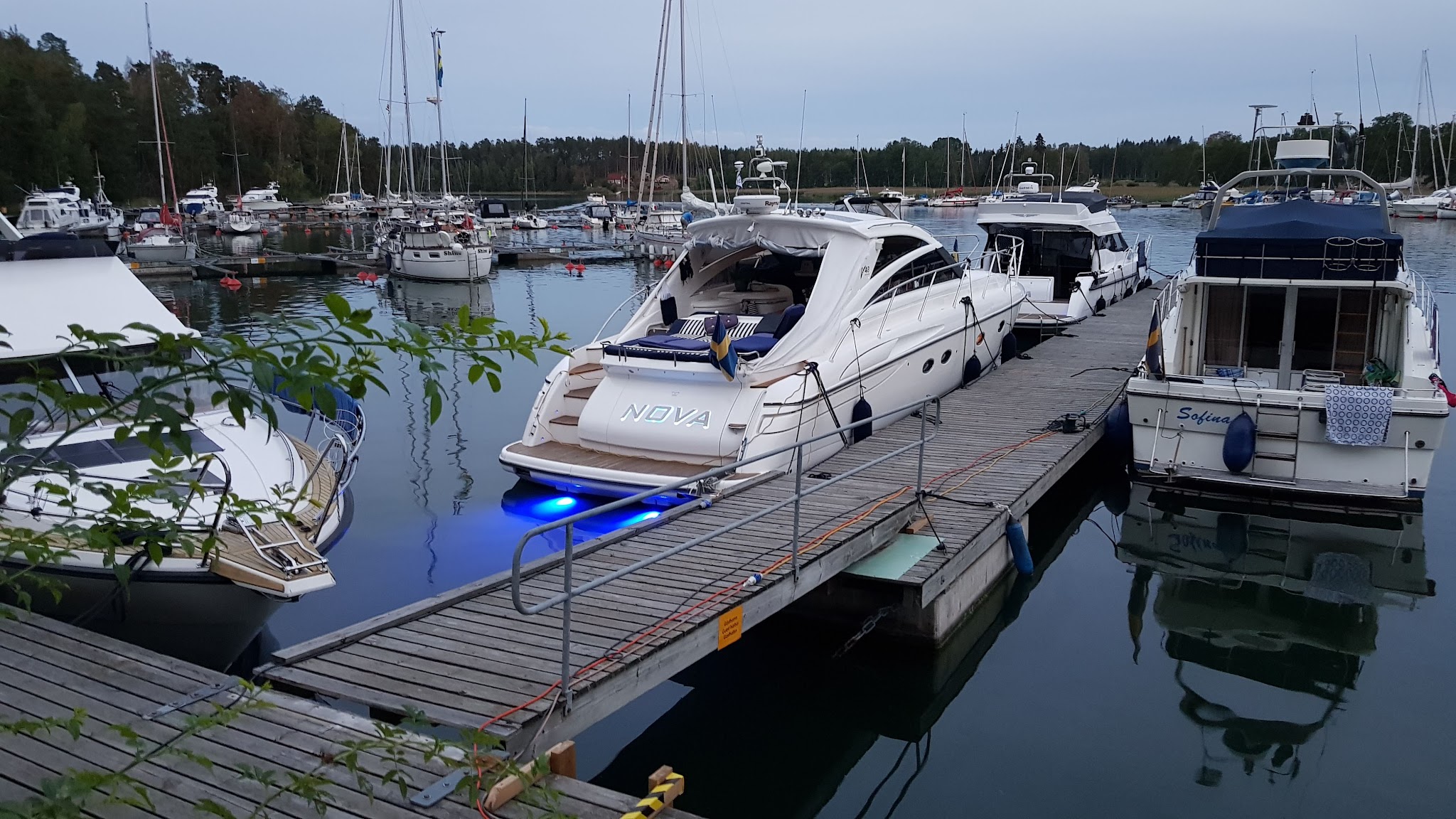 Weiße Yacht „NOVA“ mit blauem Unterwasserlicht an Holzsteg; ruhiger Yachthafen mit weiteren Booten im Abendlicht.