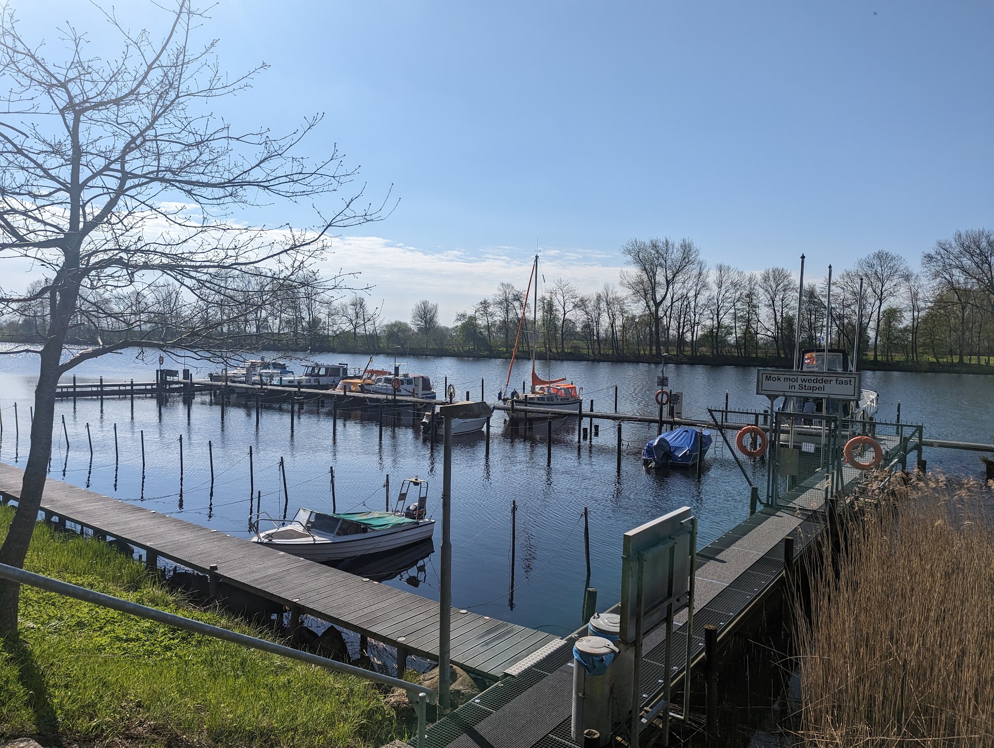 Holzstege mit kleinen Motor- und Segelbooten an ruhigem Fluss; klare Frühlingssonne, kahler Uferbaumbestand