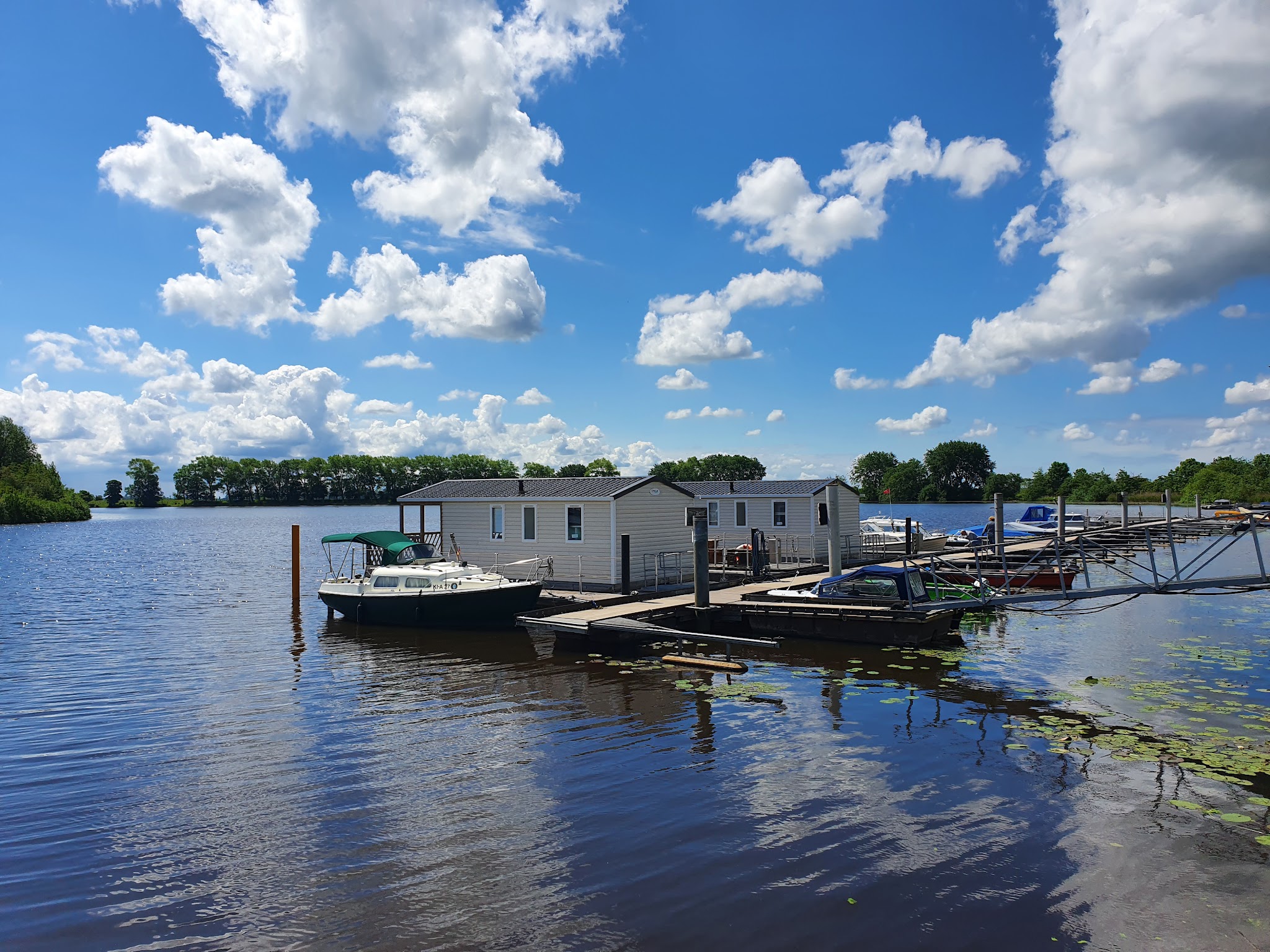 Kleiner Yachthafen mit Hausboot, anliegenden Motorbooten und Seerosen auf ruhigem See unter blauem Himmel mit Wolken.