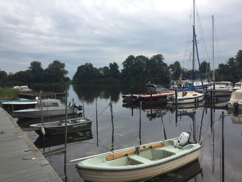 Kleine Marina; Segel- und Motorboote liegen am Steg, spiegeln sich im ruhigen Wasser vor Bäumen und grauem Himmel.