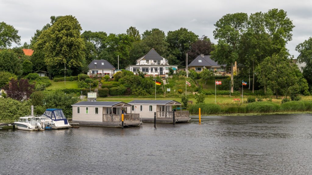 Zwei festgemachte Hausboote vor begrüntem Ufer mit Einfamilienhäusern, Bootssteg und wehenden Flaggen
