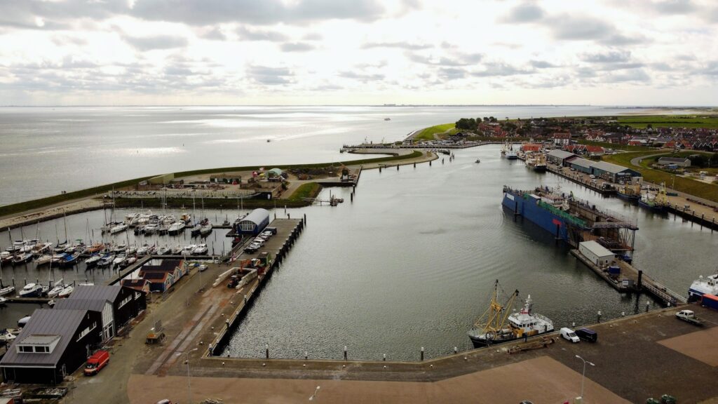 Luftaufnahme eines Küstenhafens mit Segelbooten, Werftdock und angrenzender Ortschaft unter bewölktem Himmel.