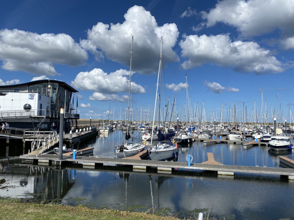Marina mit zahlreichen Segelbooten an Stegen, links moderner Hafenbau; ruhiges Wasser spiegelt blauen Himmel mit Wolken