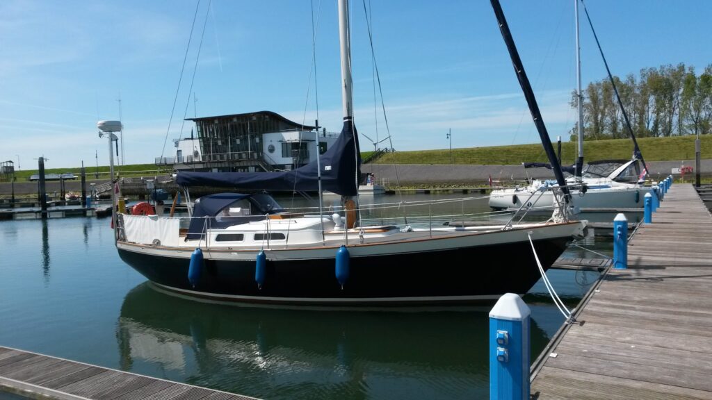 Segelboot liegt an Holzsteg im ruhigen Yachthafen bei Sonnenschein, modernes Hafenhaus im Hintergrund.