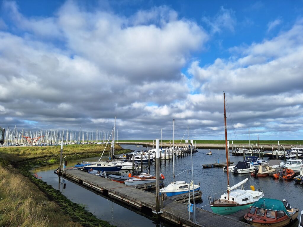 Kleiner Yachthafen: Segel- und Motorboote an Holzstegen, dahinter Deich, darüber blauer Himmel mit Wolken.
