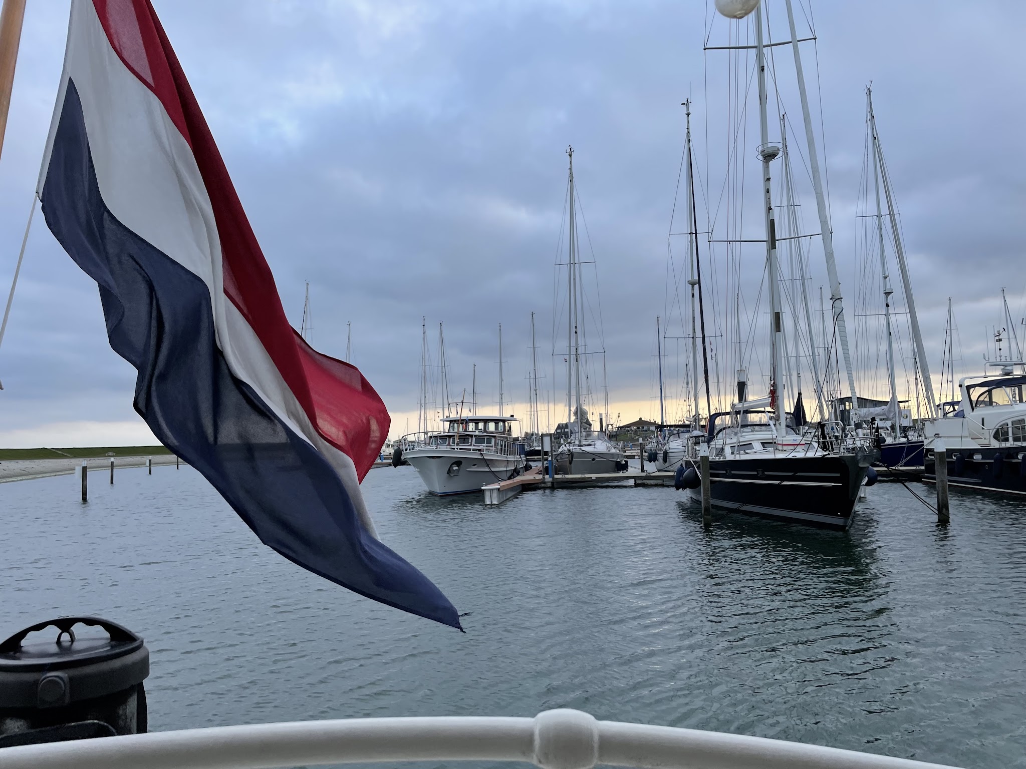 Niederländische Flagge weht vor ruhigem Jachthafen mit angelegten Segelbooten unter bewölktem Abendhimmel.