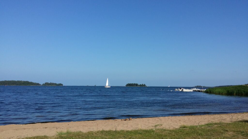 Sandstrand mit Gras, blaues Wasser, Segelboot und Motorboote vor kleinen bewaldeten Inseln unter wolkenlosem Himmel.