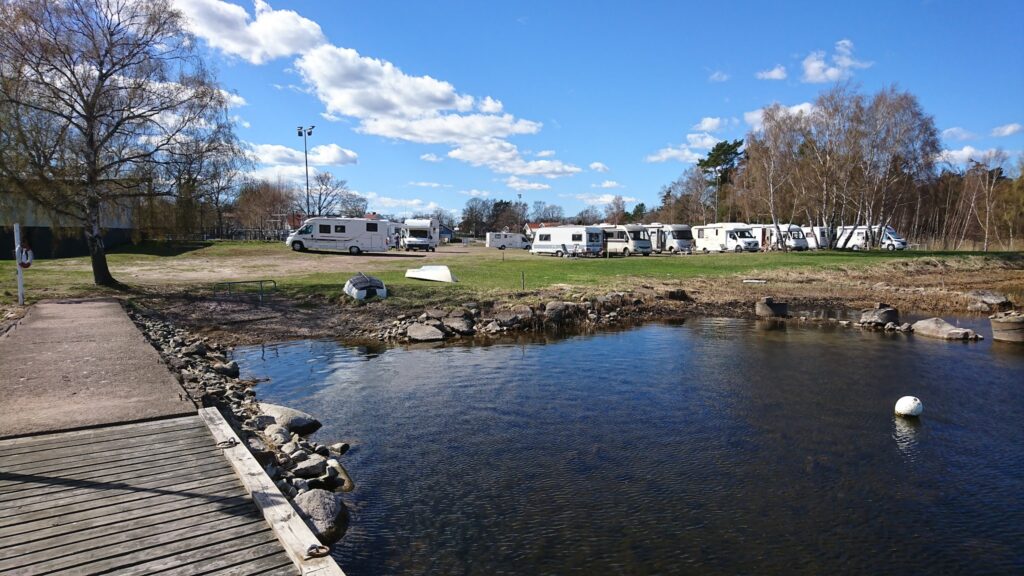 RV-Campsite an ruhigem See; links Holzsteg, gegenüber auf Wiese mehrere Wohnmobile unter blauem Himmel.