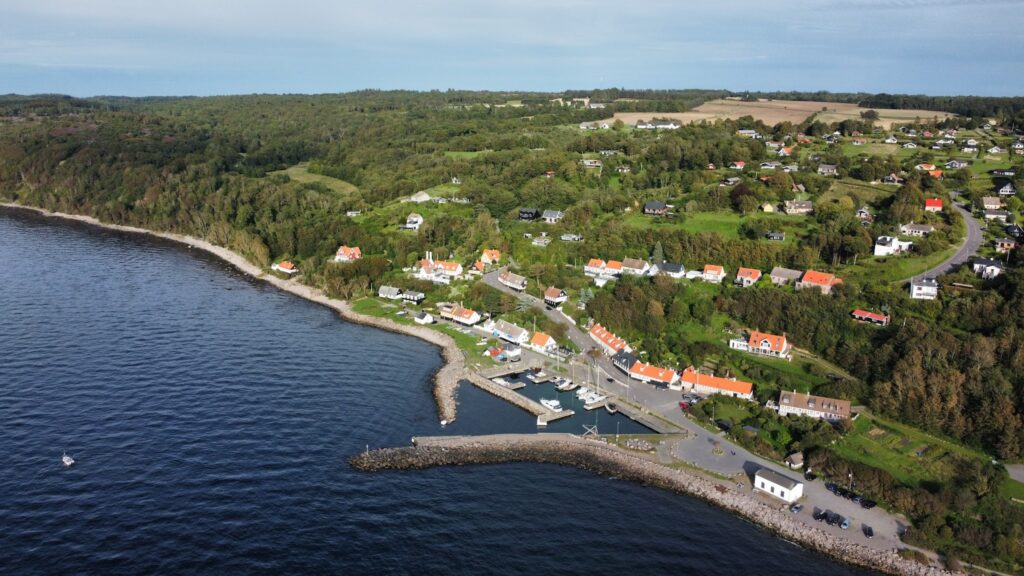 Luftaufnahme eines kleinen Küstendorfs mit roten Dächern, kleinem Yachthafen und dicht bewaldeten Hügeln am Meer.