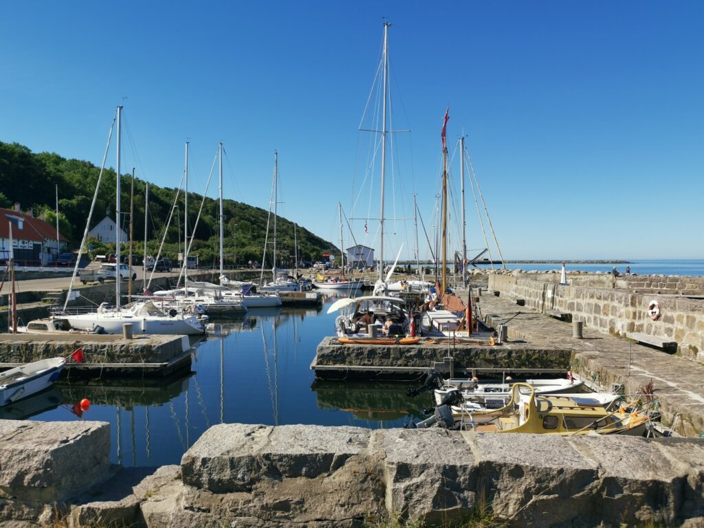 Kleiner Hafen mit festgemachten Segelbooten, ruhiges Wasser, Steinpier, grüner Hügel und klarer blauer Himmel.